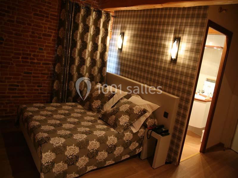 Chambre avec lit double, murs en briques et à motifs, éclairage mural et vue partielle sur une salle de bain.