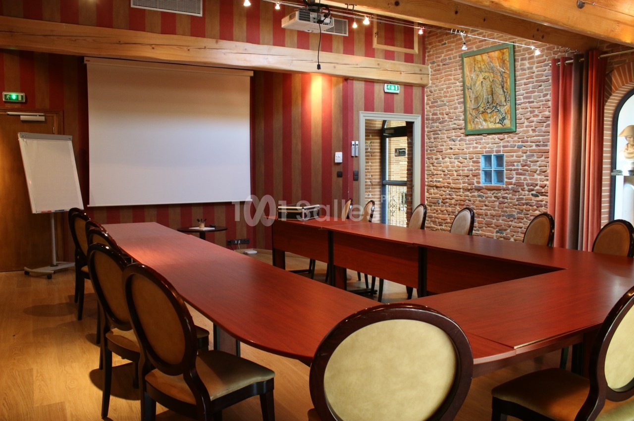 Salle de réunion avec tables en bois disposées en U, chaises rembourrées, écran de projection et mur en briques apparentes.