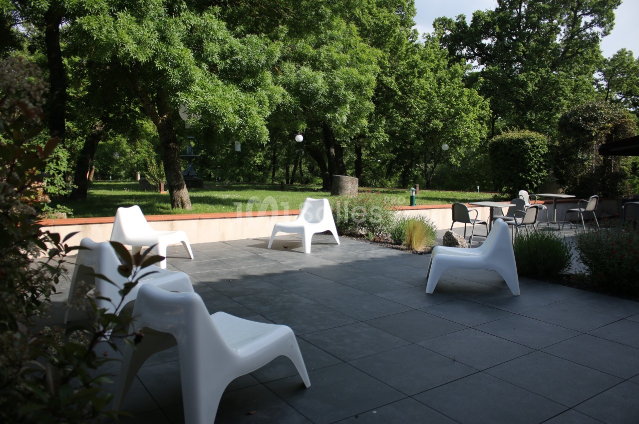 Terrasse avec chaises blanches modernes, entourée de verdure et d'arbres, sous une lumière naturelle.