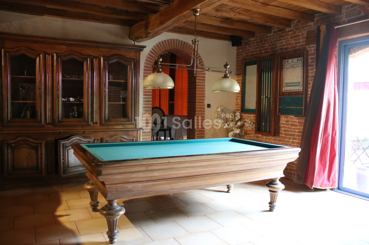 Table de billard en bois avec tapis vert dans une pièce au style rustique, avec poutres apparentes et murs en briques.