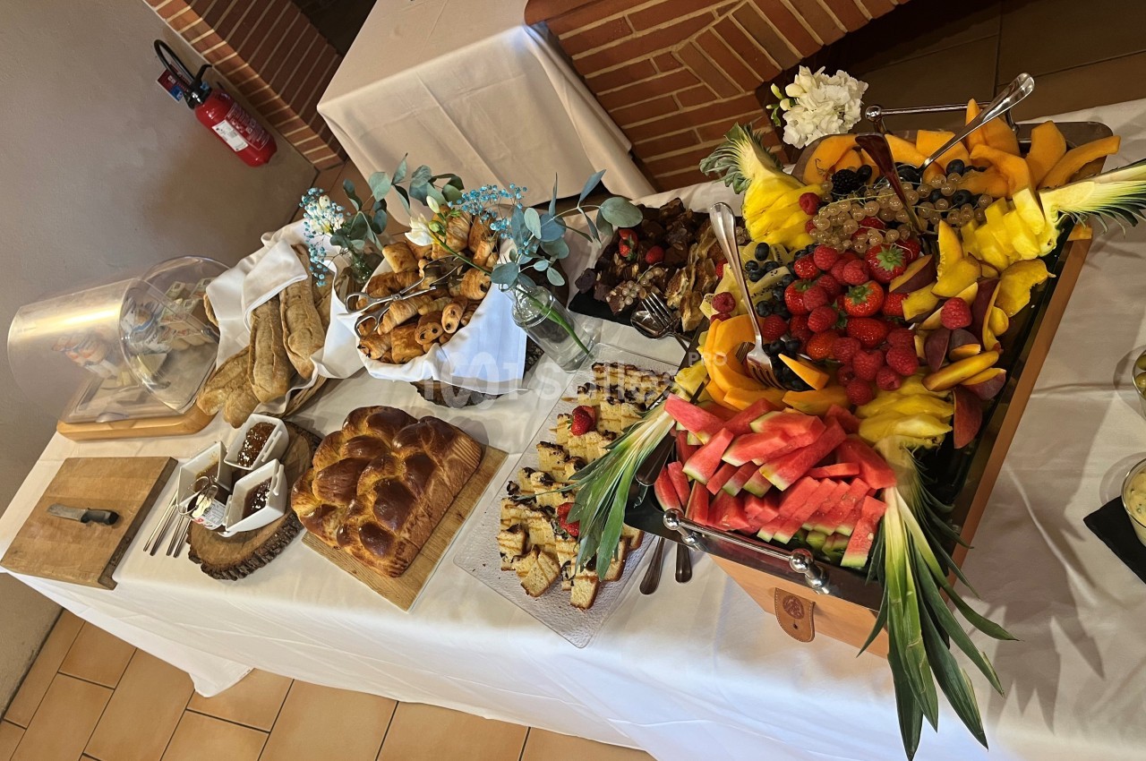 Buffet varié avec fruits frais, viennoiseries, pains, et desserts disposés sur une table blanche.