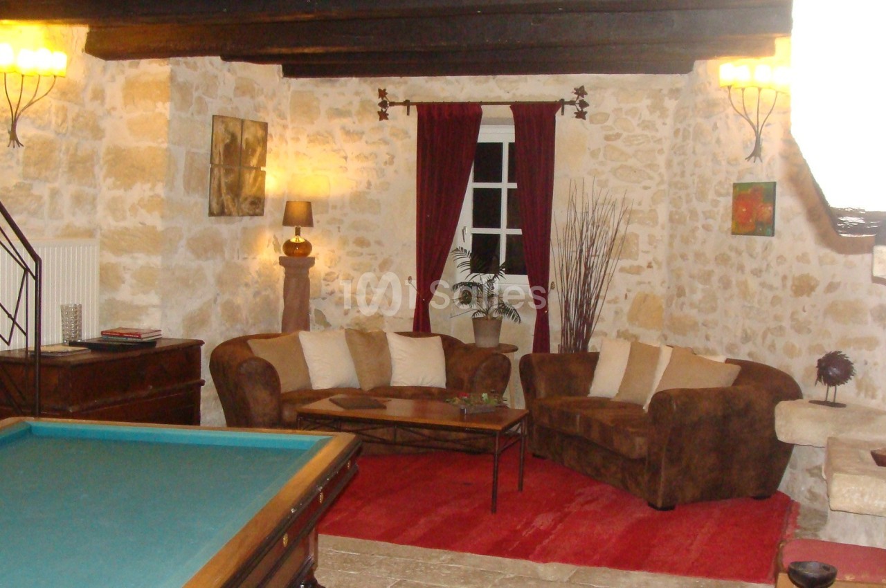 Salon rustique avec murs en pierre, canapé en velours marron, table basse, tapis rouge et table de billard au premier plan.