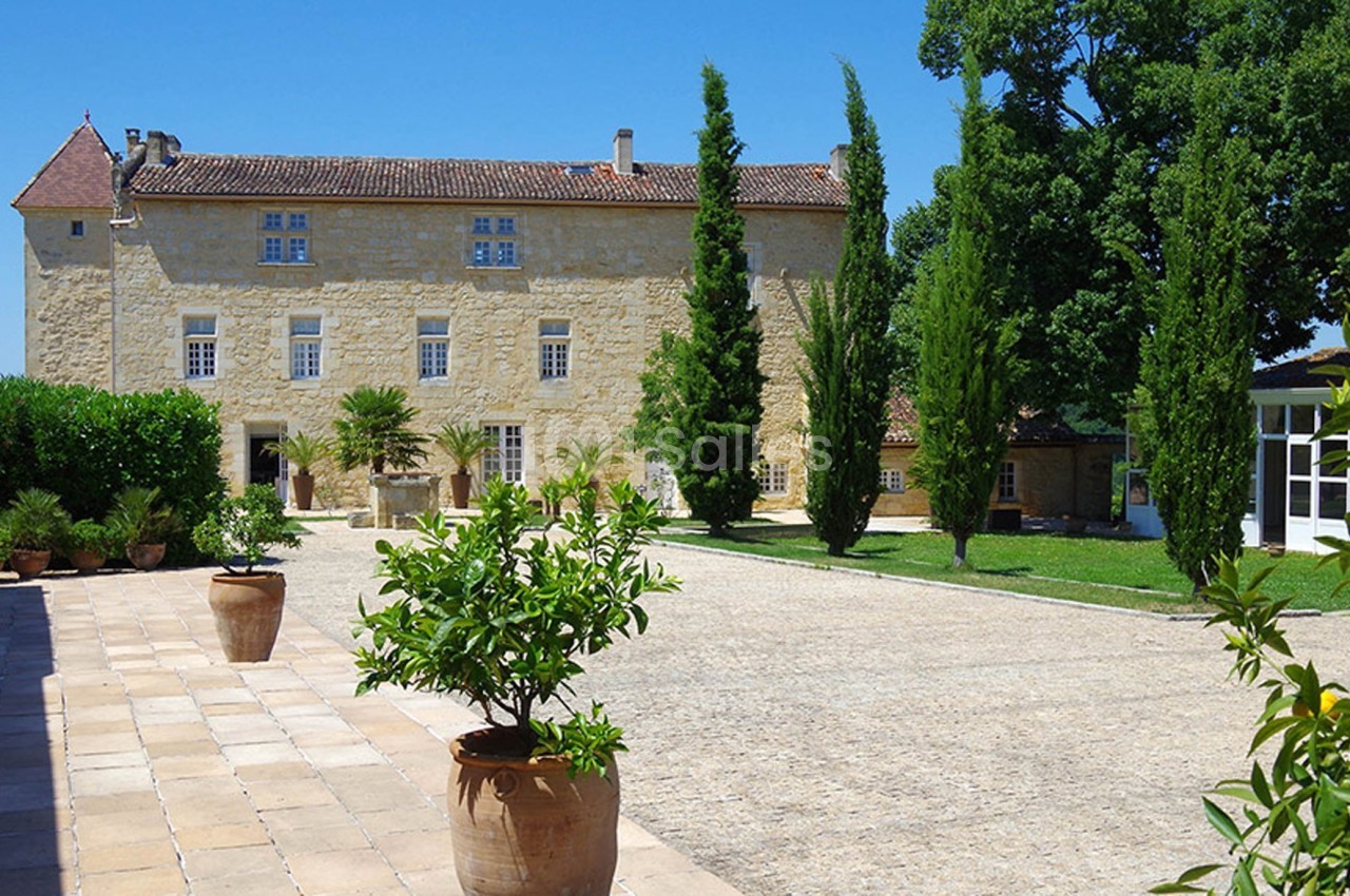 Cour pavée avec des pots de plantes, bordée d'un bâtiment en pierre à plusieurs étages et d'arbres verts.