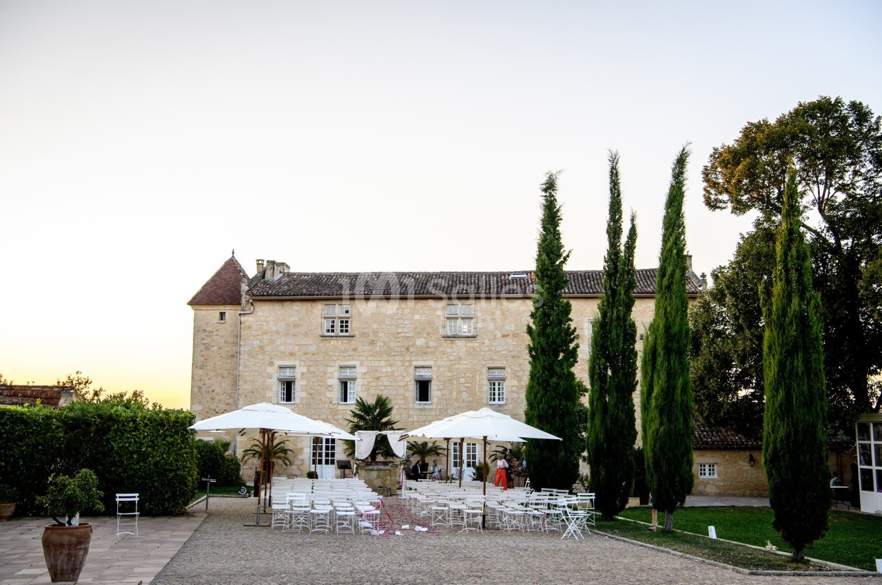 Cour d'un château en pierre avec des tables et chaises blanches disposées sous des parasols, entourée de cyprès.
