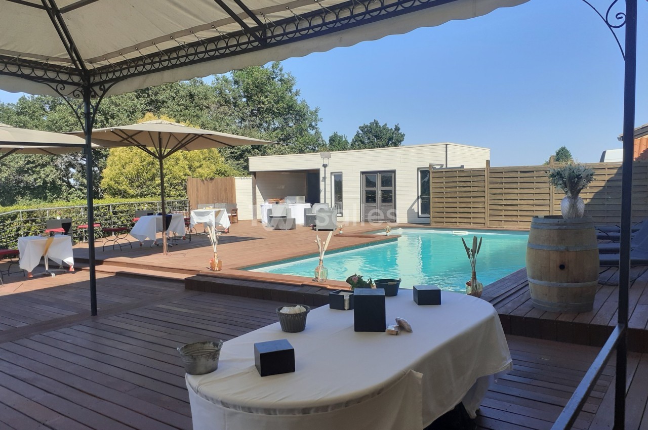 Terrasse en bois avec tables dressées, piscine extérieure et pergola, entourée de végétation et d'une clôture.
