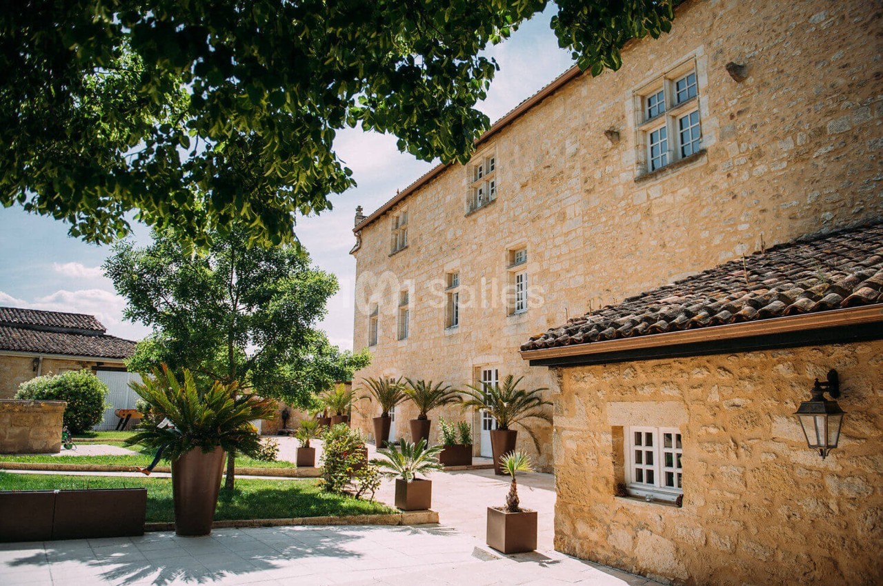 Façade en pierre d'un bâtiment ancien entouré d'arbres, de plantes en pots et d'une cour pavée.