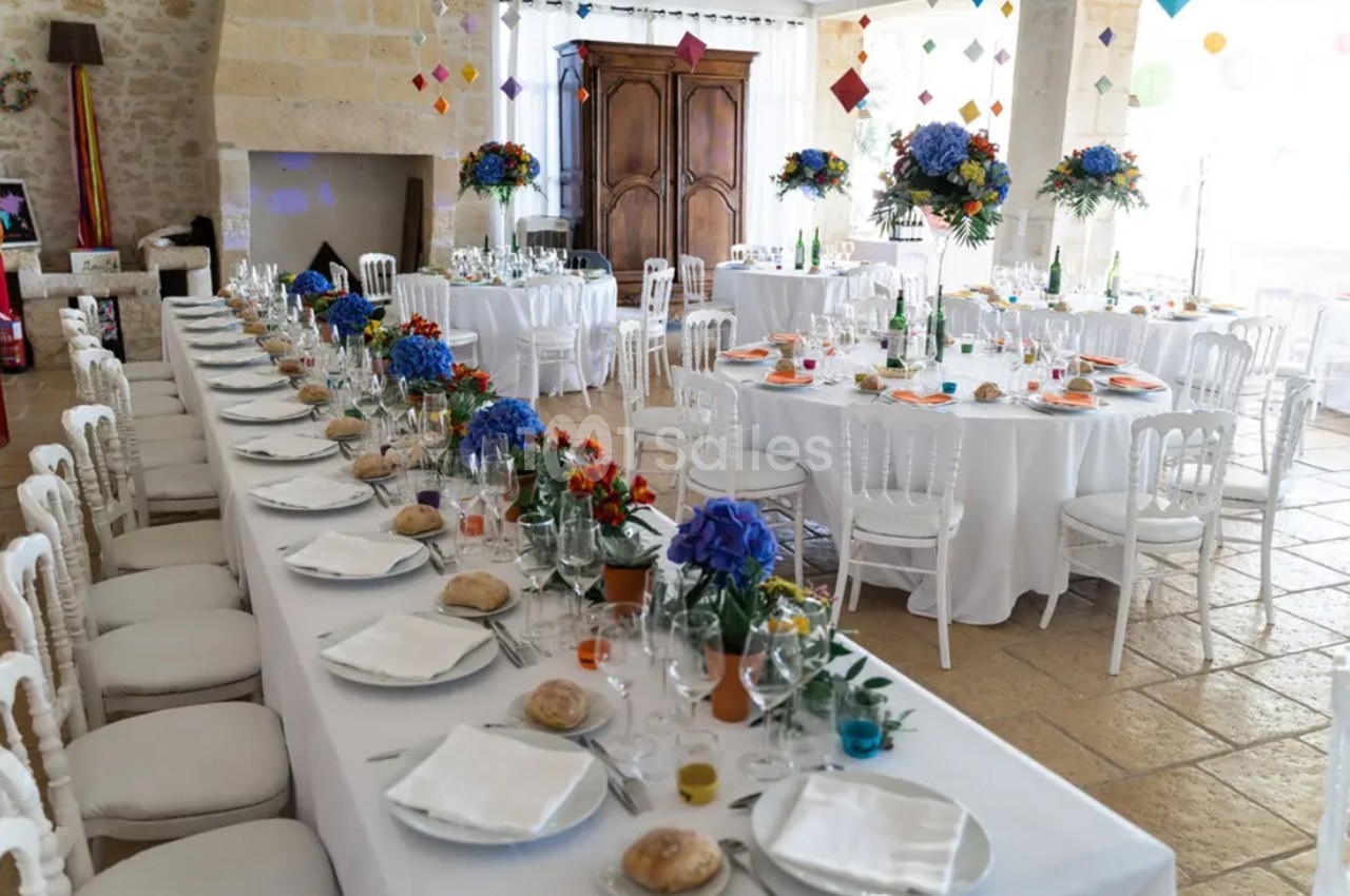 Salle de réception décorée pour un événement, avec tables dressées, nappes blanches et arrangements floraux colorés.