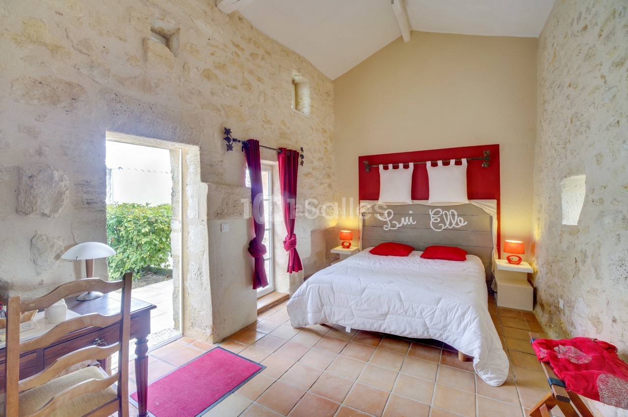 Chambre lumineuse avec murs en pierre, lit double, rideaux rouges et accès à une terrasse extérieure.