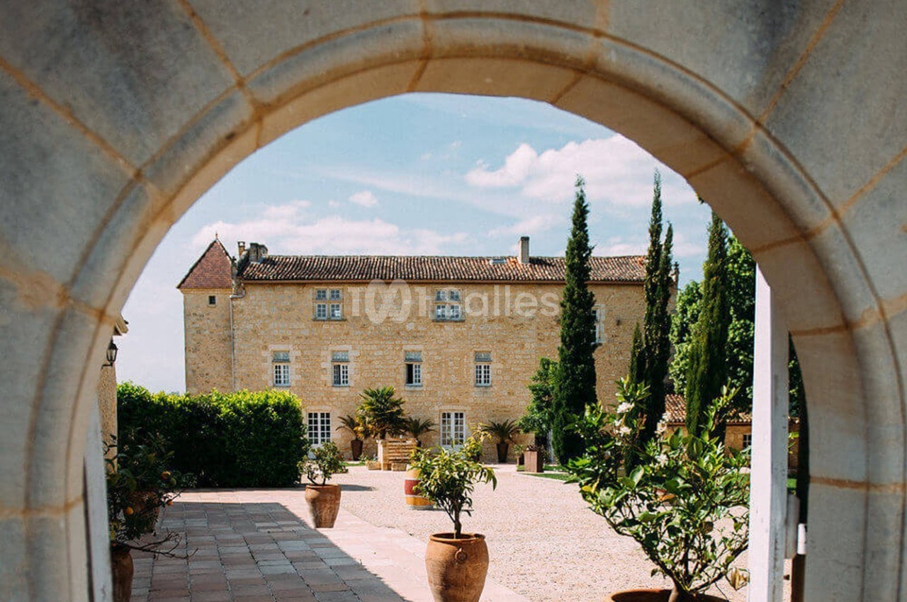 Vue d'un château en pierre entouré de cyprès et de plantes en pot, encadré par une arche en pierre.