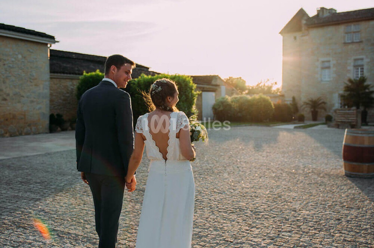 Un couple en tenue de mariage marche main dans la main dans une cour pavée au coucher du soleil.