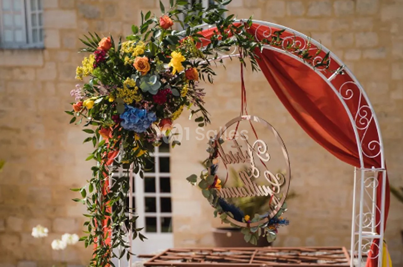 Arche décorative en métal blanc ornée de fleurs colorées et de rubans rouges, devant un mur en pierre beige.