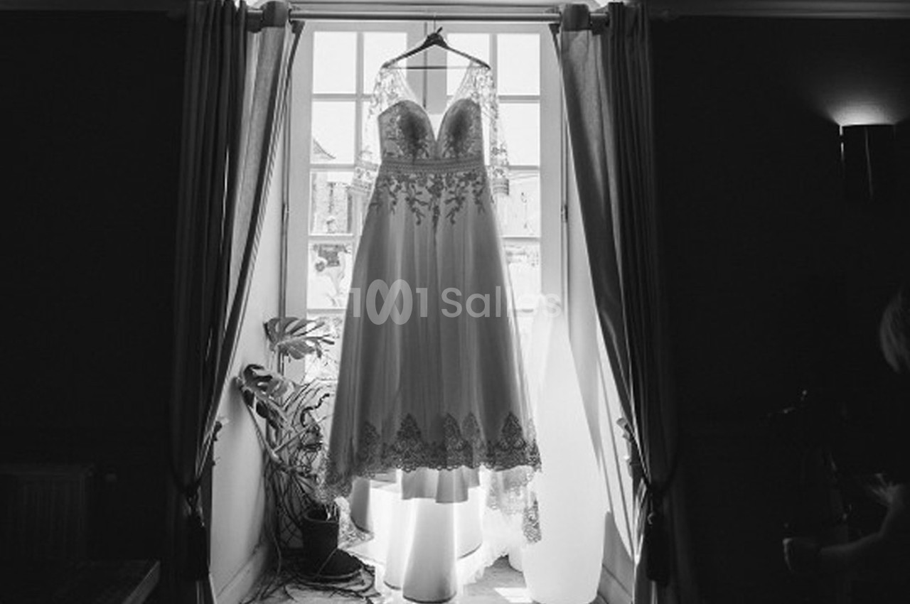 Robe de mariée en dentelle suspendue devant une fenêtre lumineuse avec des rideaux ouverts.