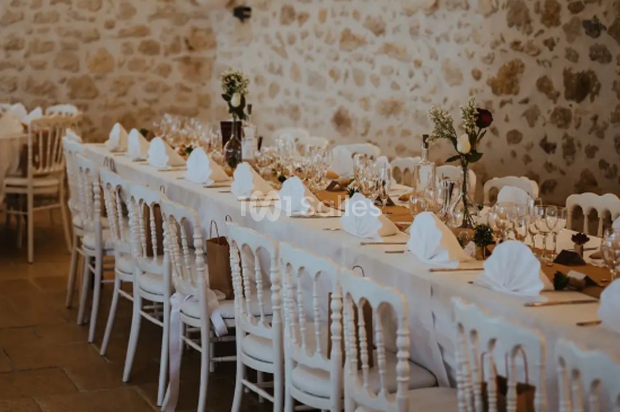 Grande table de banquet décorée avec nappes blanches, serviettes pliées et vaisselle, dans une salle en pierre.