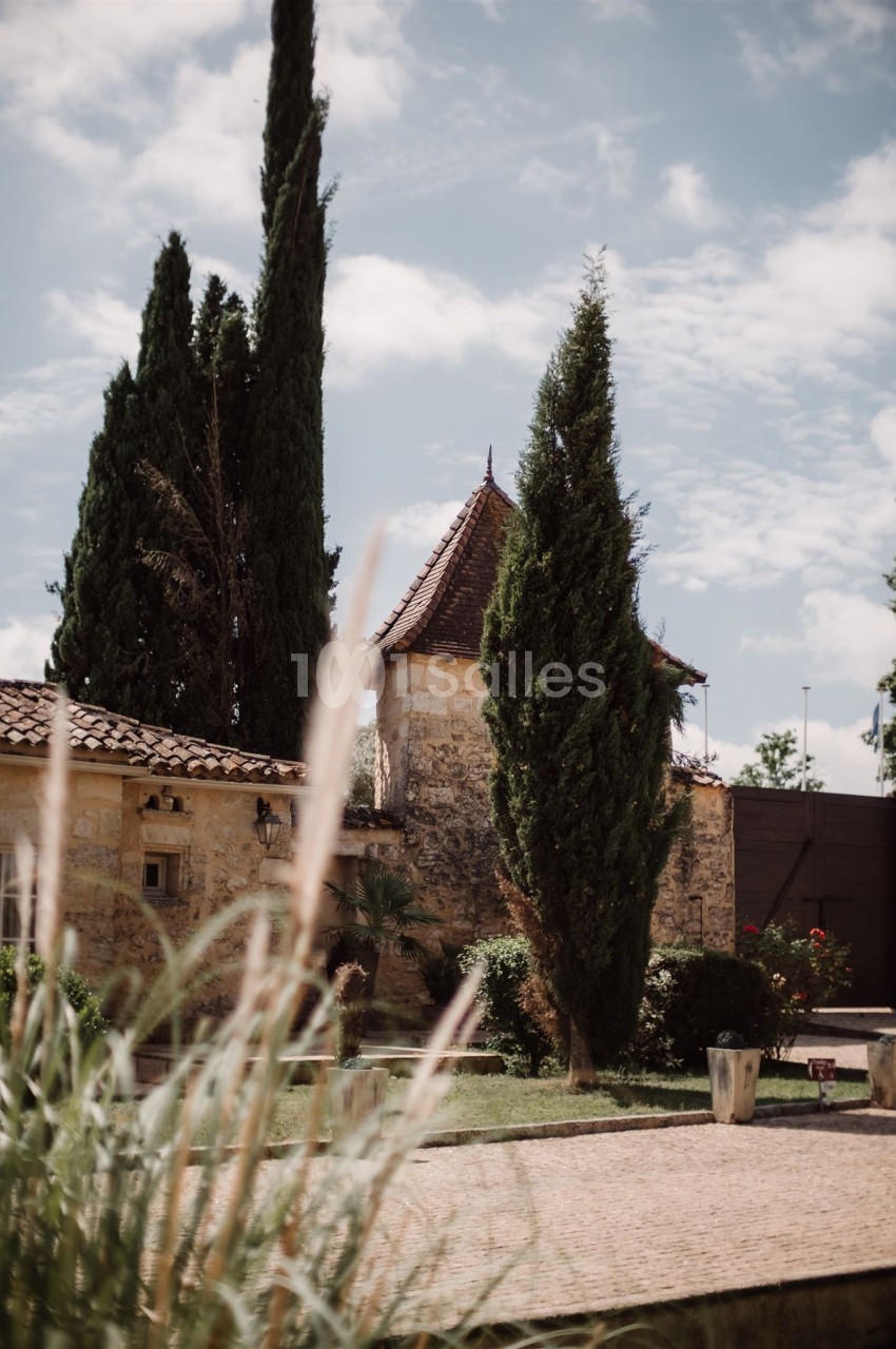 Vue d'un jardin avec des cyprès, un bâtiment en pierre aux tuiles rouges et un ciel partiellement nuageux.