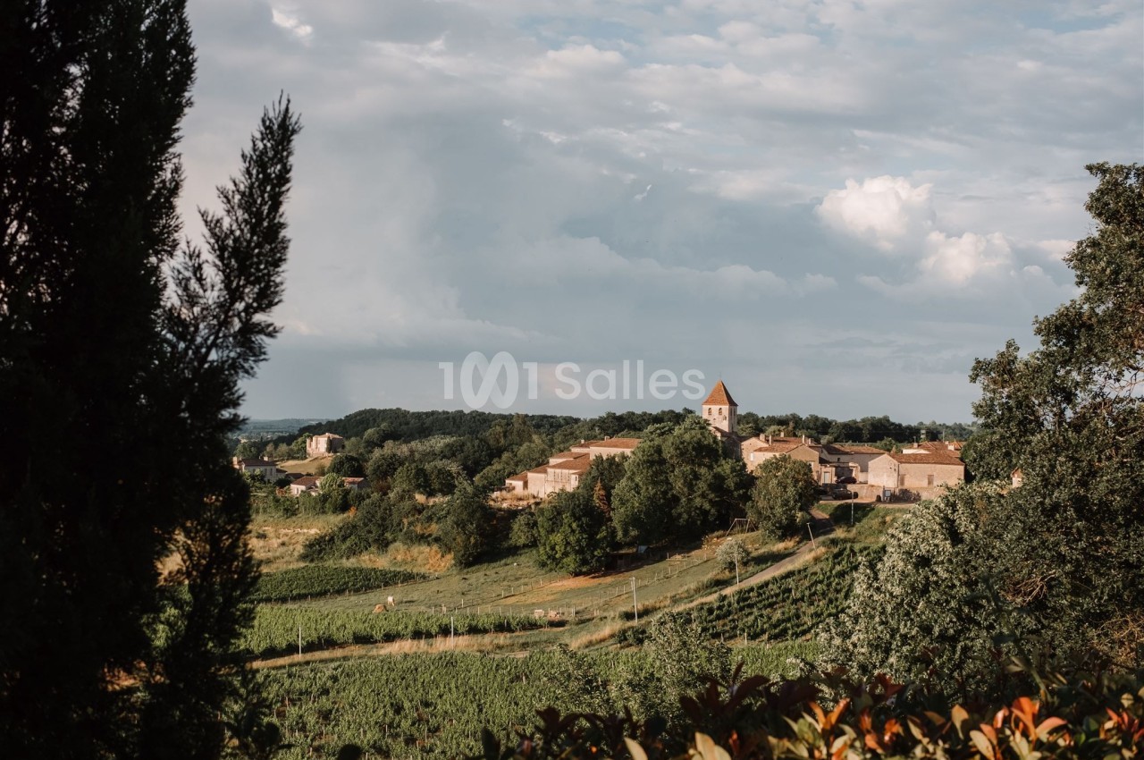 Village rural avec église au clocher pointu, entouré de champs verdoyants et d'arbres sous un ciel partiellement nuageux.