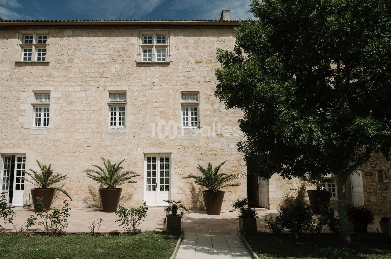 Façade en pierre d'une maison avec plusieurs fenêtres, terrasse ornée de plantes en pots et un arbre à droite.