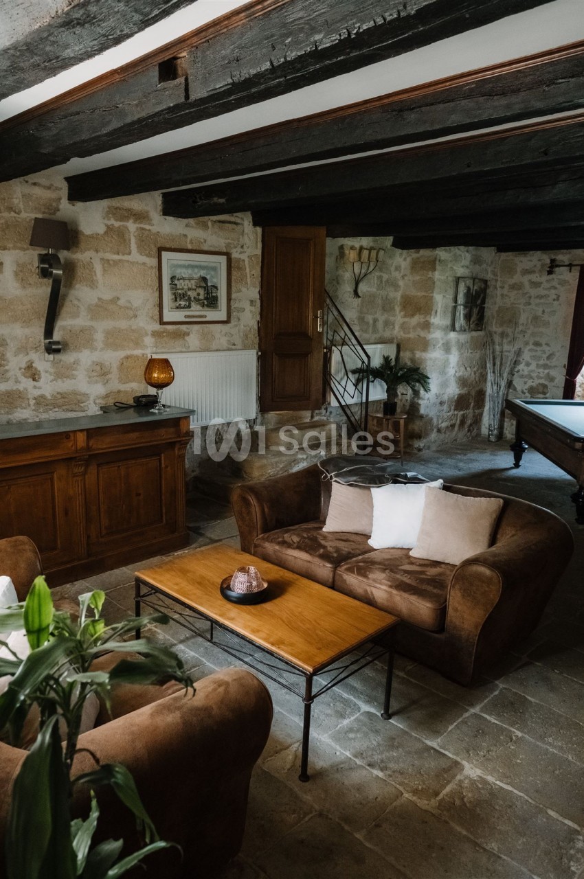Salon rustique avec murs en pierre, poutres apparentes, canapé en velours brun, table basse en bois et décoration…