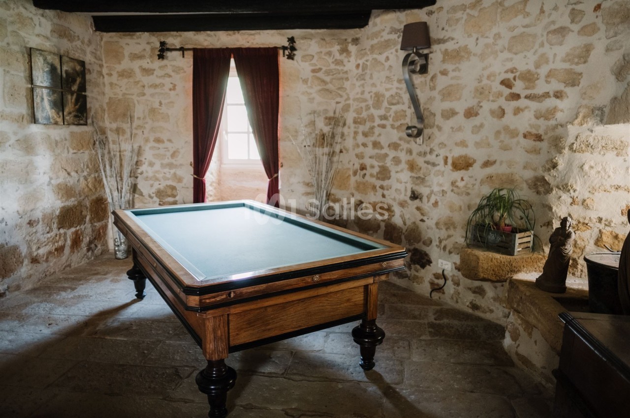 Table de billard en bois dans une pièce en pierre avec un mur éclairé par une fenêtre encadrée de rideaux rouges.