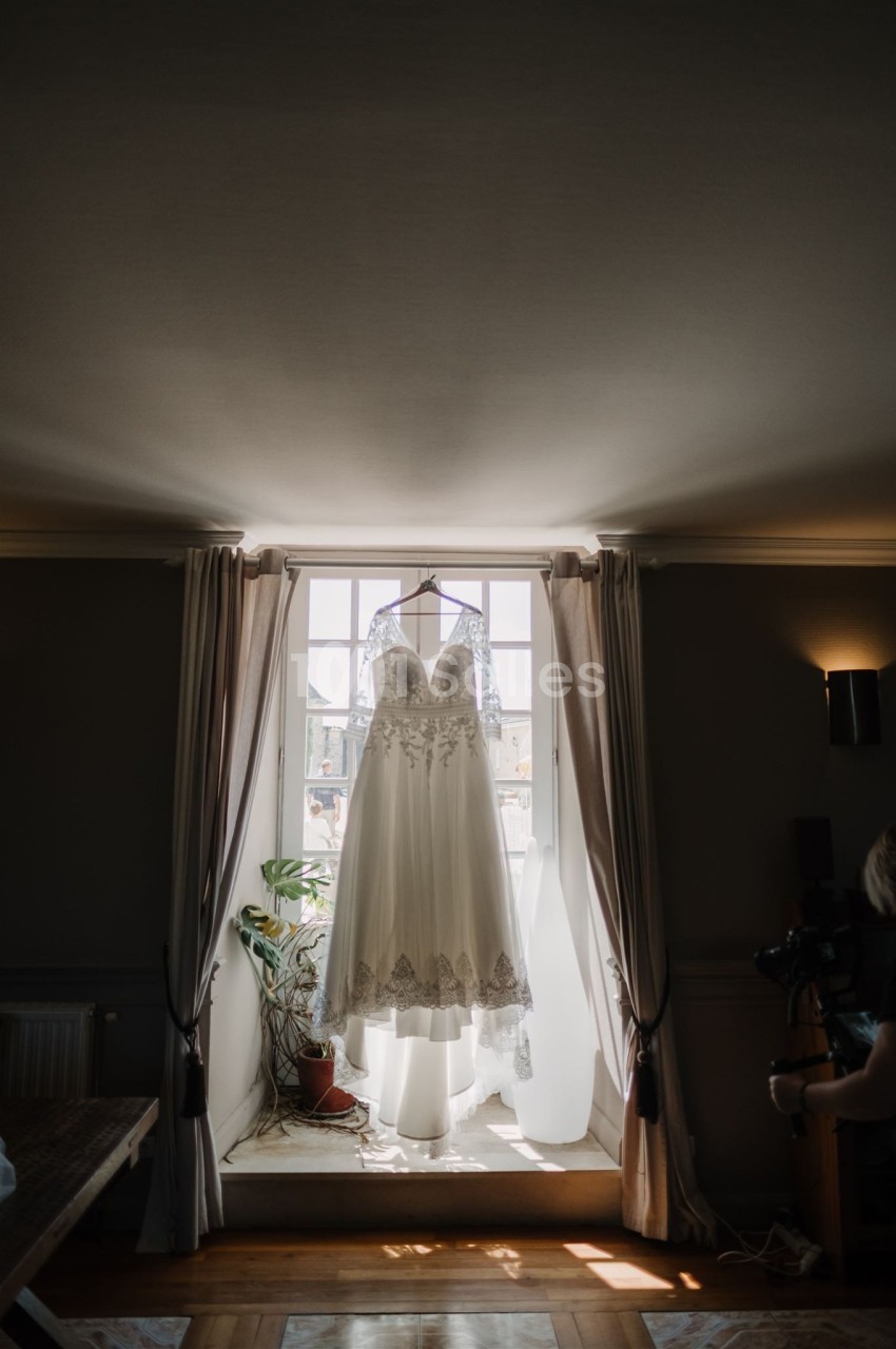 Robe de mariée blanche en dentelle suspendue devant une fenêtre lumineuse avec des rideaux ouverts.