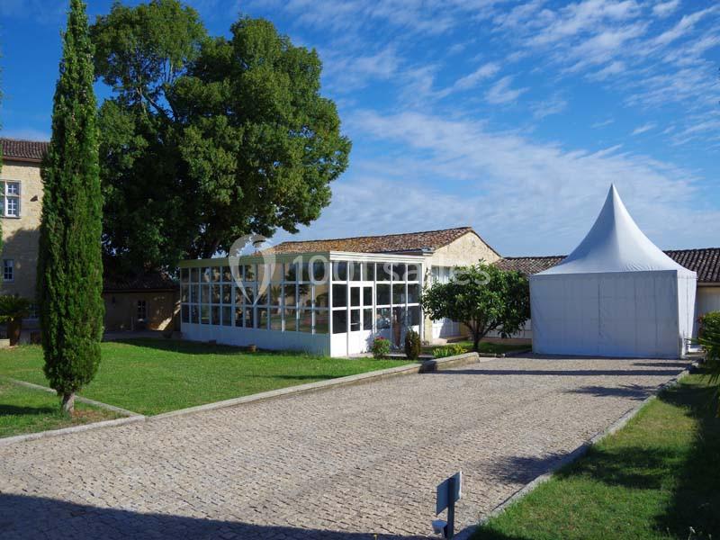Bâtiment avec une façade vitrée, une tente blanche et un chemin pavé, entourés de verdure sous un ciel bleu.