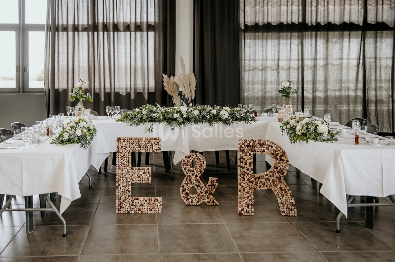 Décoration de table de mariage avec nappes blanches, fleurs, pampas et initiales ’E & R’ en bouchons de liège.