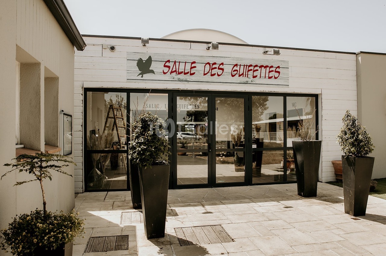 Entrée d'un bâtiment avec une enseigne ’Salle des Guifettes’, entourée de plantes en pots sur une terrasse pavée.