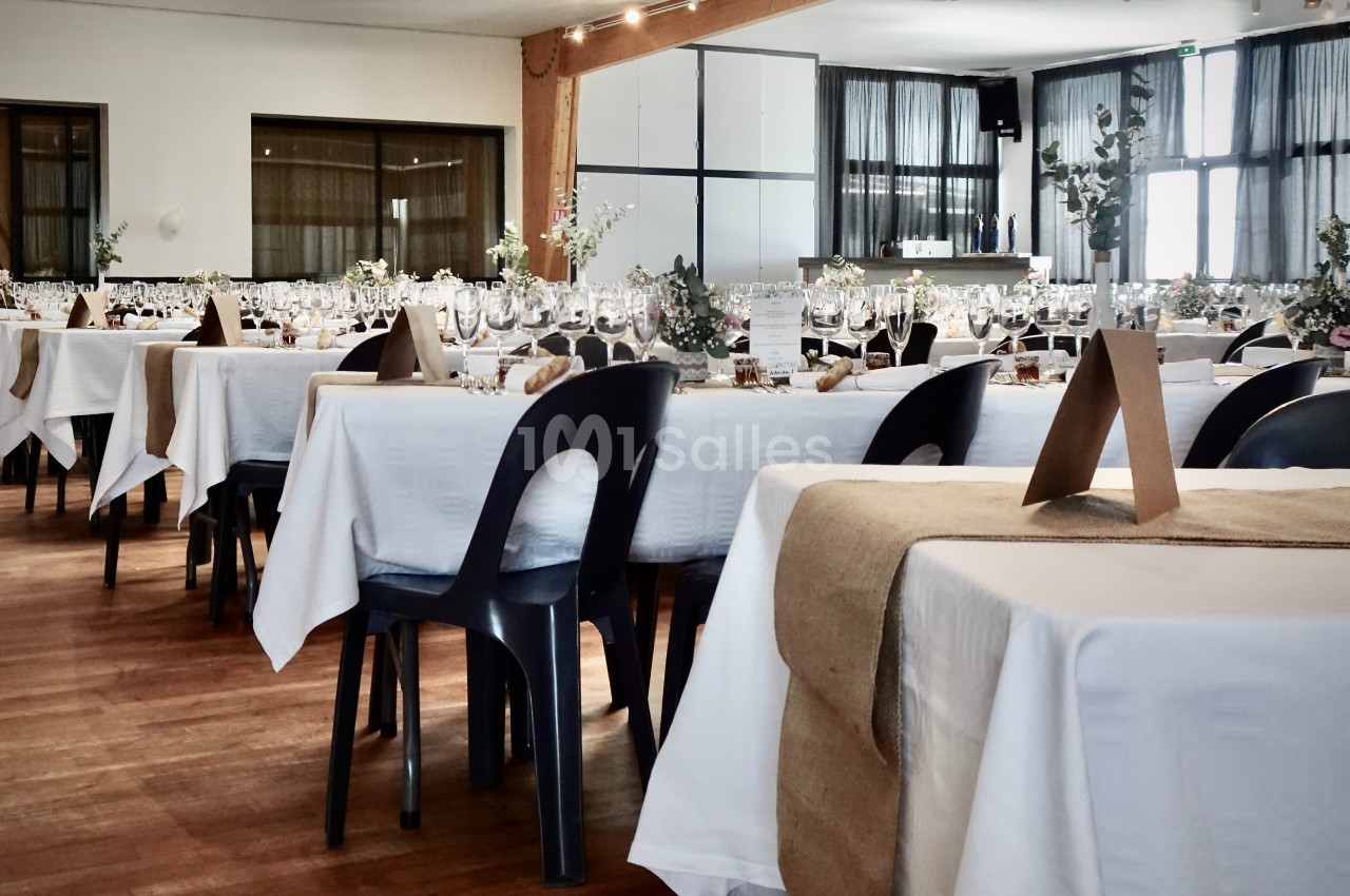 Salle de réception décorée avec des tables dressées, nappes blanches, chemins de table en jute et chaises noires.