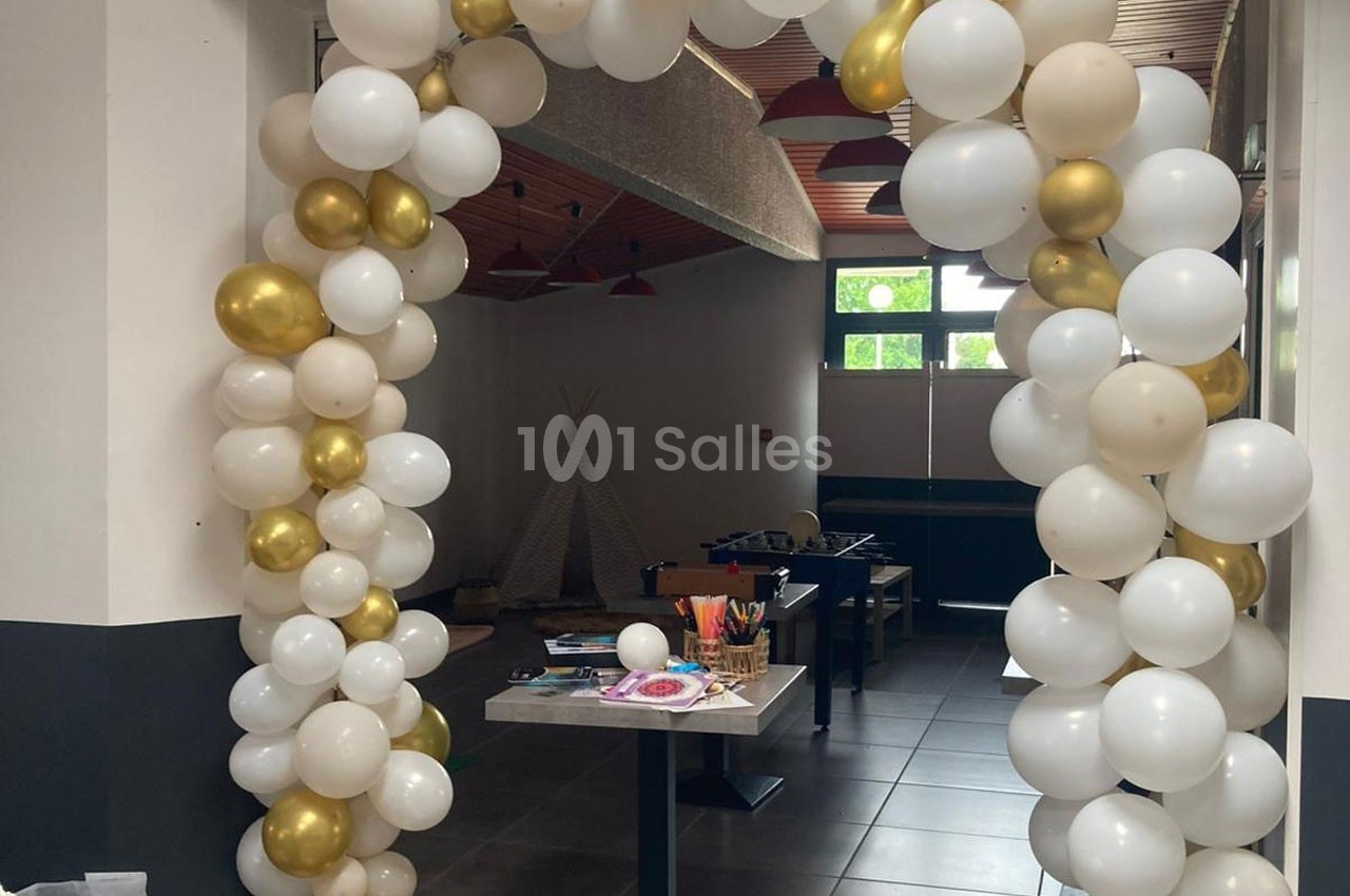Arche décorative en ballons blancs et dorés dans une pièce avec des tables et des jeux en arrière-plan.