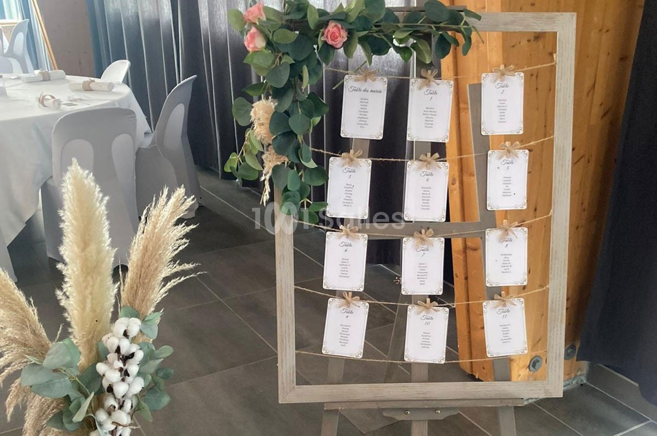 Plan de table de mariage affiché sur un cadre en bois décoré de fleurs et feuillages, avec des tables dressées en arrière…