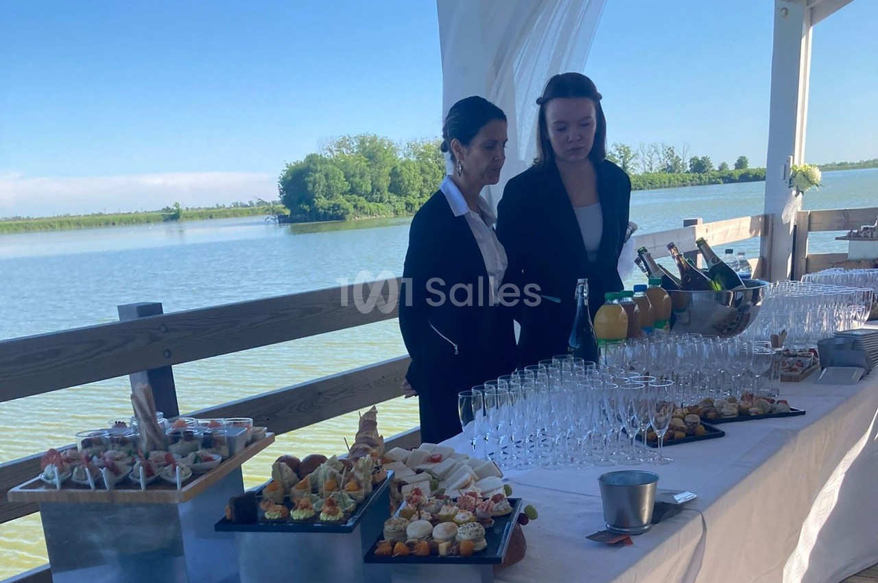 Deux personnes devant une table dressée avec des amuse-bouches, des boissons et des verres, près d'un plan d'eau.