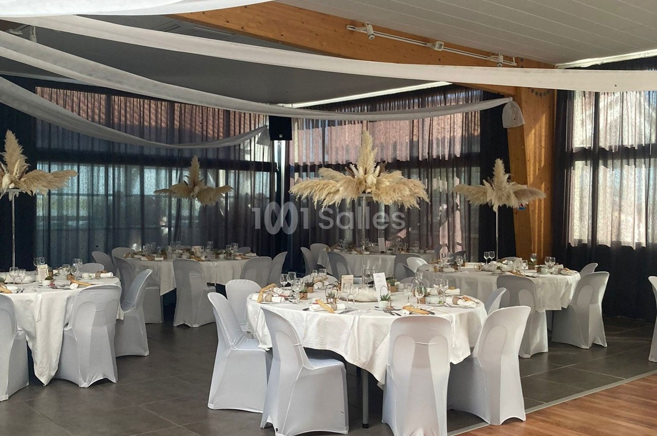 Salle de réception décorée avec des tables rondes, nappes blanches, chaises houssées et centres de table en pampas.