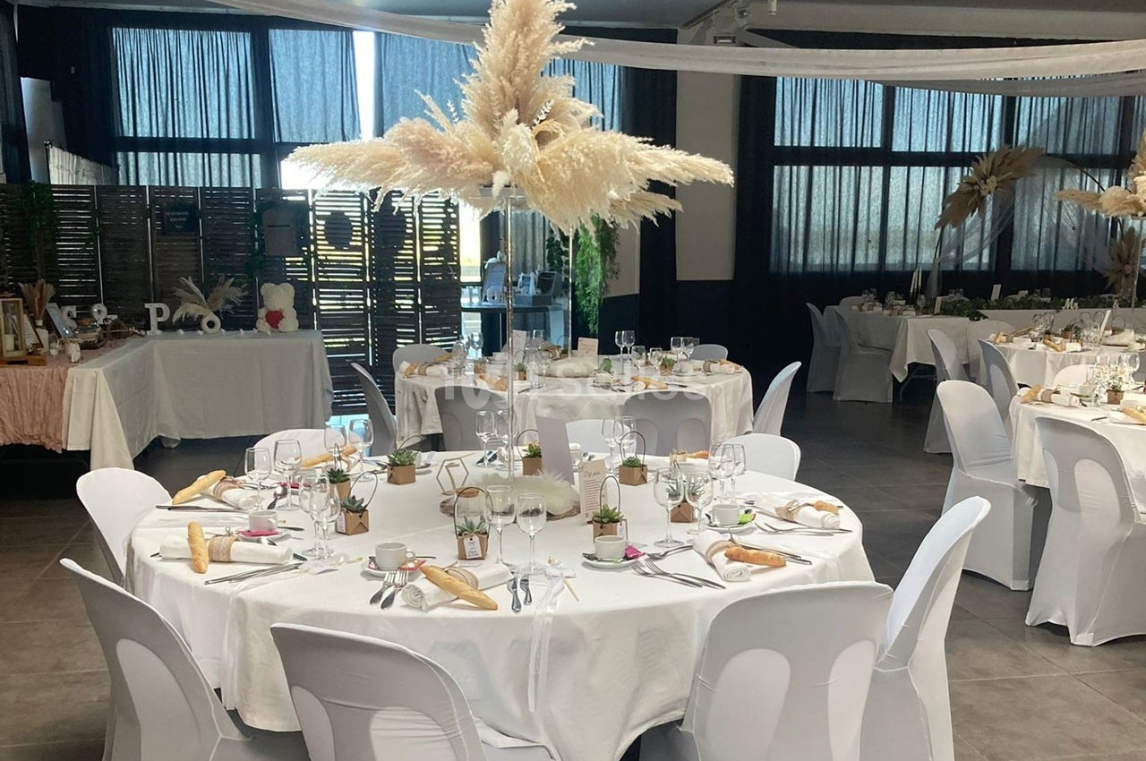 Salle de réception décorée avec des tables rondes dressées, nappes blanches et centre de table en herbes de pampa.