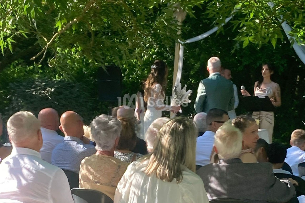 Cérémonie de mariage en plein air avec des invités assis, un couple devant un officiant sous un arbre.