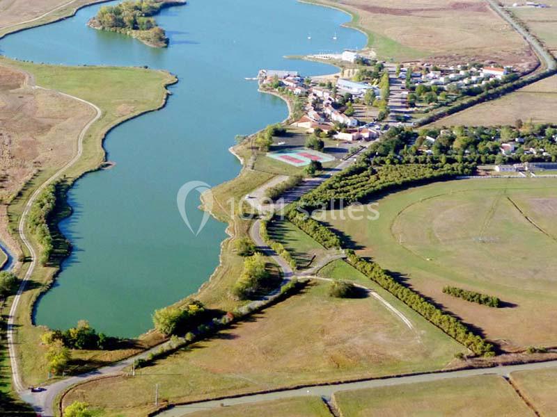 Vue aérienne d'un lac entouré de verdure, avec un village et des infrastructures sportives à proximité.