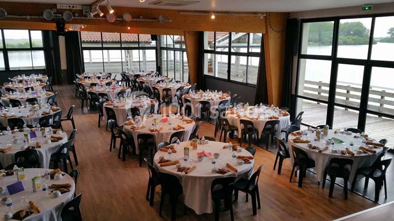 Salle de réception lumineuse avec des tables rondes dressées pour un repas, vue sur un extérieur avec plan d'eau.
