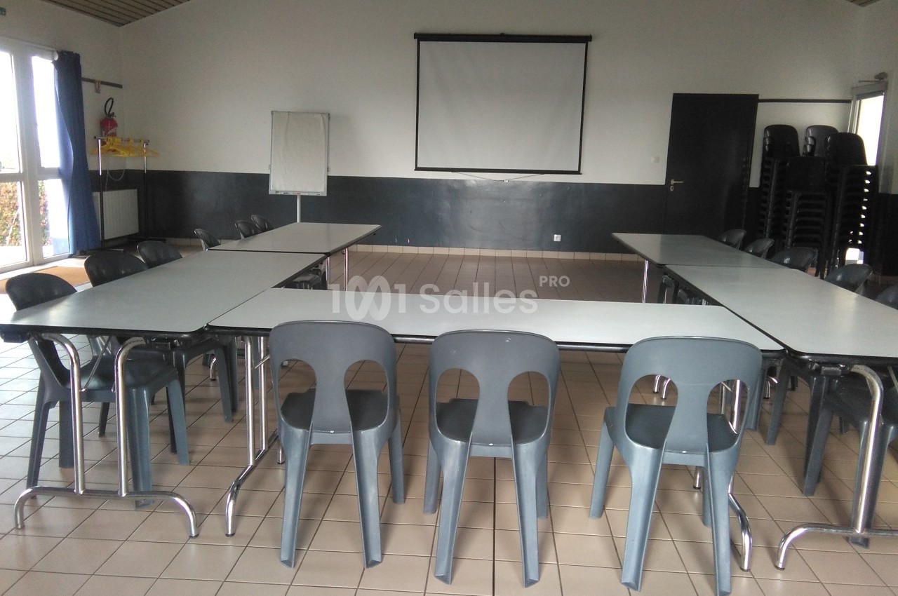 Salle de réunion vide avec tables disposées en U, chaises en plastique gris et écran de projection au fond.