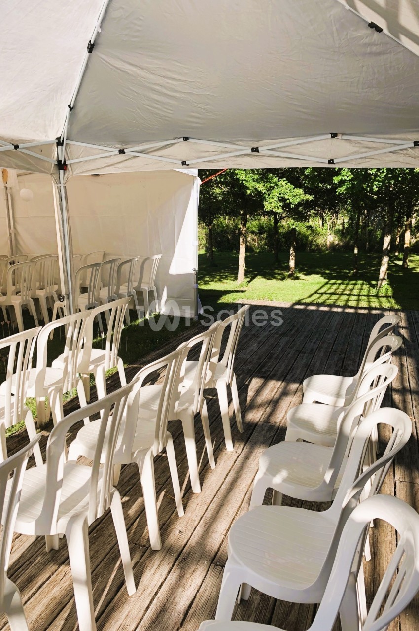 Chaises blanches en plastique alignées sous une tente blanche sur une terrasse en bois, avec un jardin en arrière-plan.