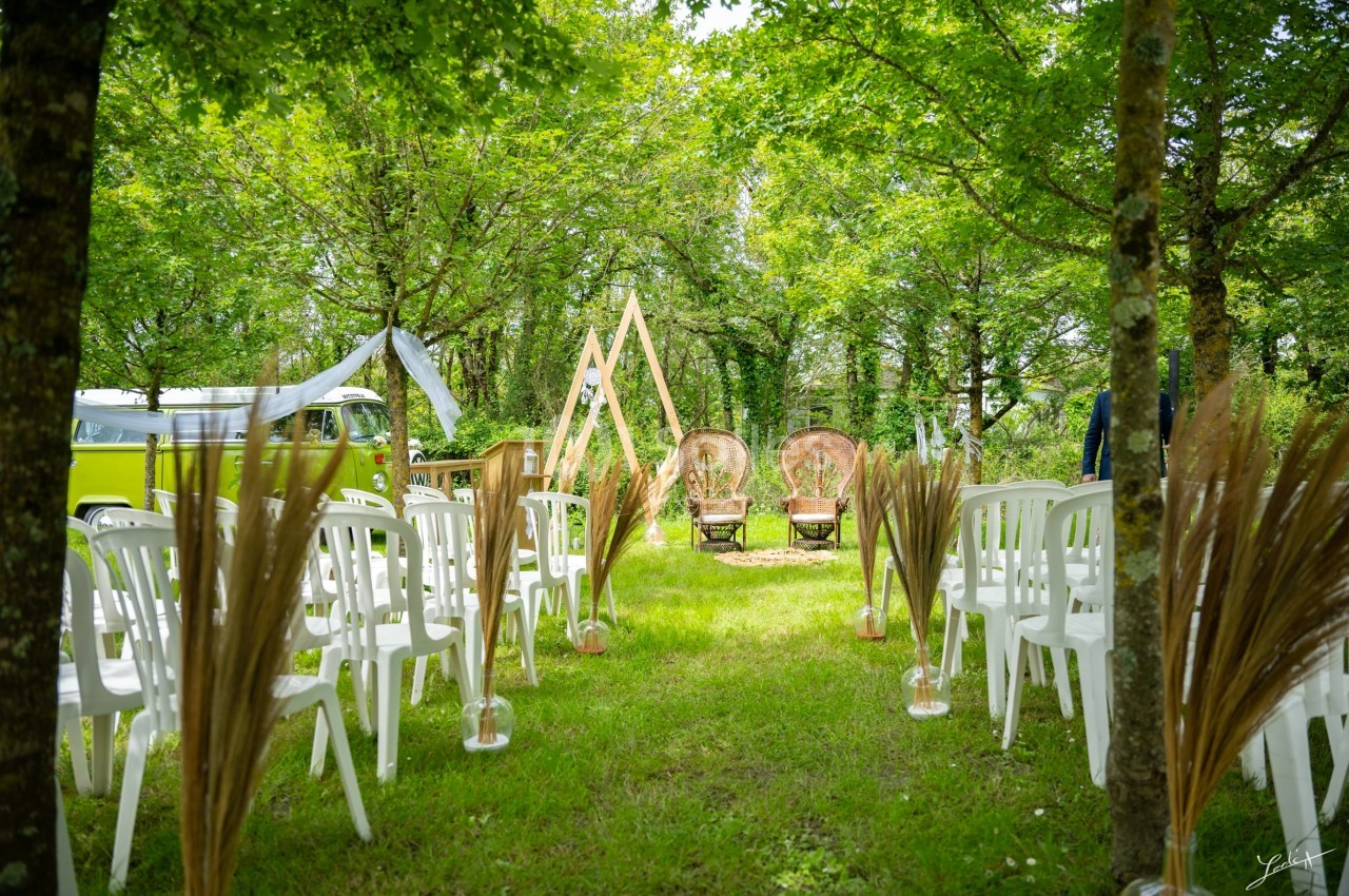 Allée de chaises blanches disposées en extérieur, menant à une arche en bois et des fauteuils sous des arbres.