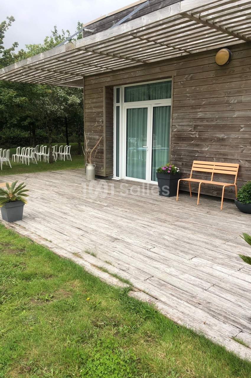 Terrasse en bois avec banc, pots de fleurs et pergola, donnant sur une pelouse et des chaises blanches en arrière-plan.