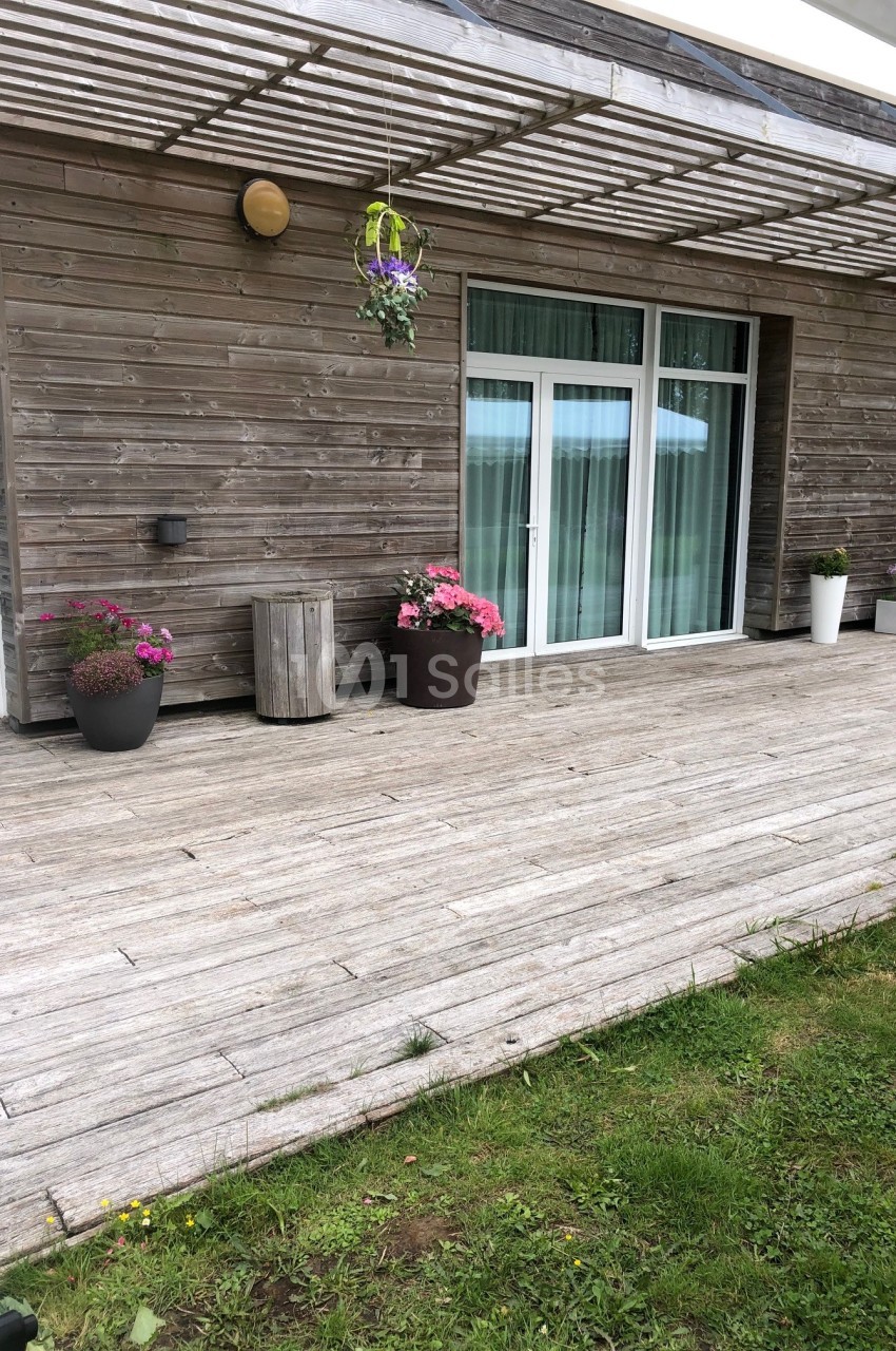 Terrasse en bois avec pots de fleurs, pergola en bois et grandes baies vitrées donnant sur l'intérieur.