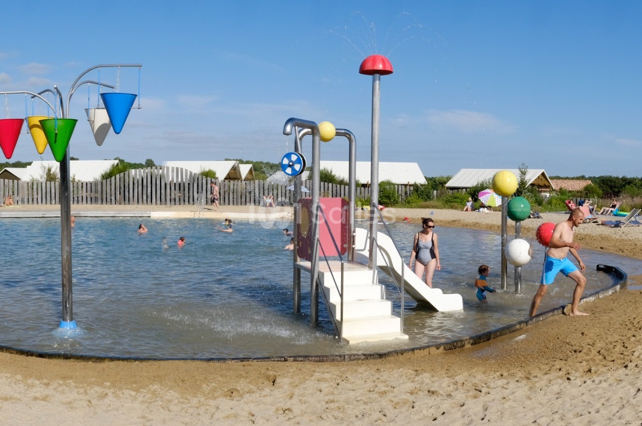 Bassin peu profond avec jeux d'eau colorés, toboggan et plage de sable, entouré de végétation et d'une clôture en bois.
