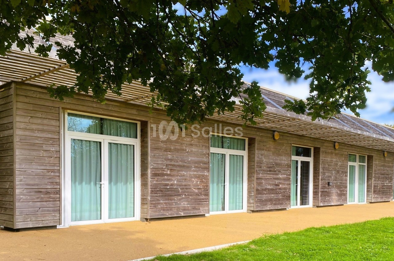Bâtiment en bois avec de grandes baies vitrées, entouré de verdure et partiellement ombragé par des arbres.