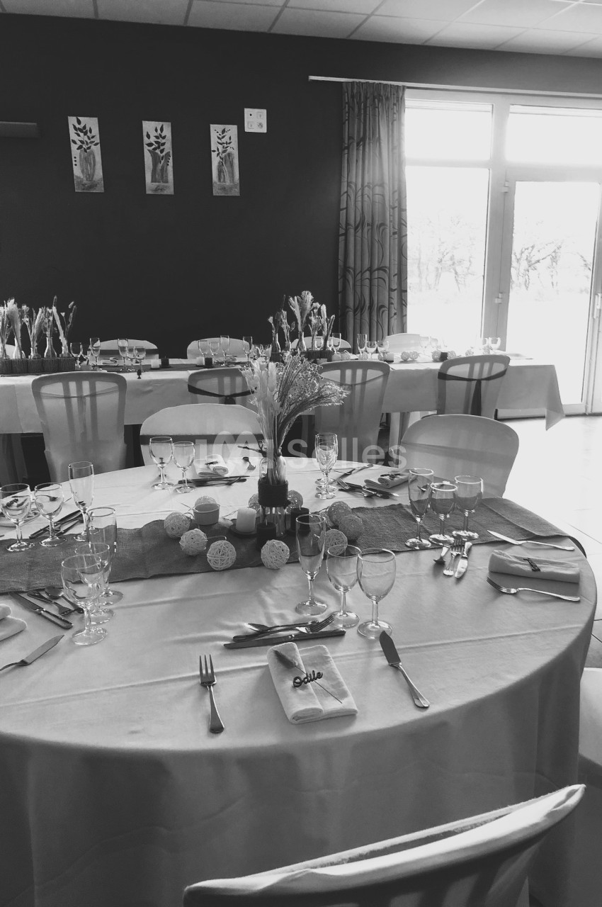 Salle de réception avec tables dressées, nappes blanches, décorations florales et lumière naturelle entrant par une baie…
