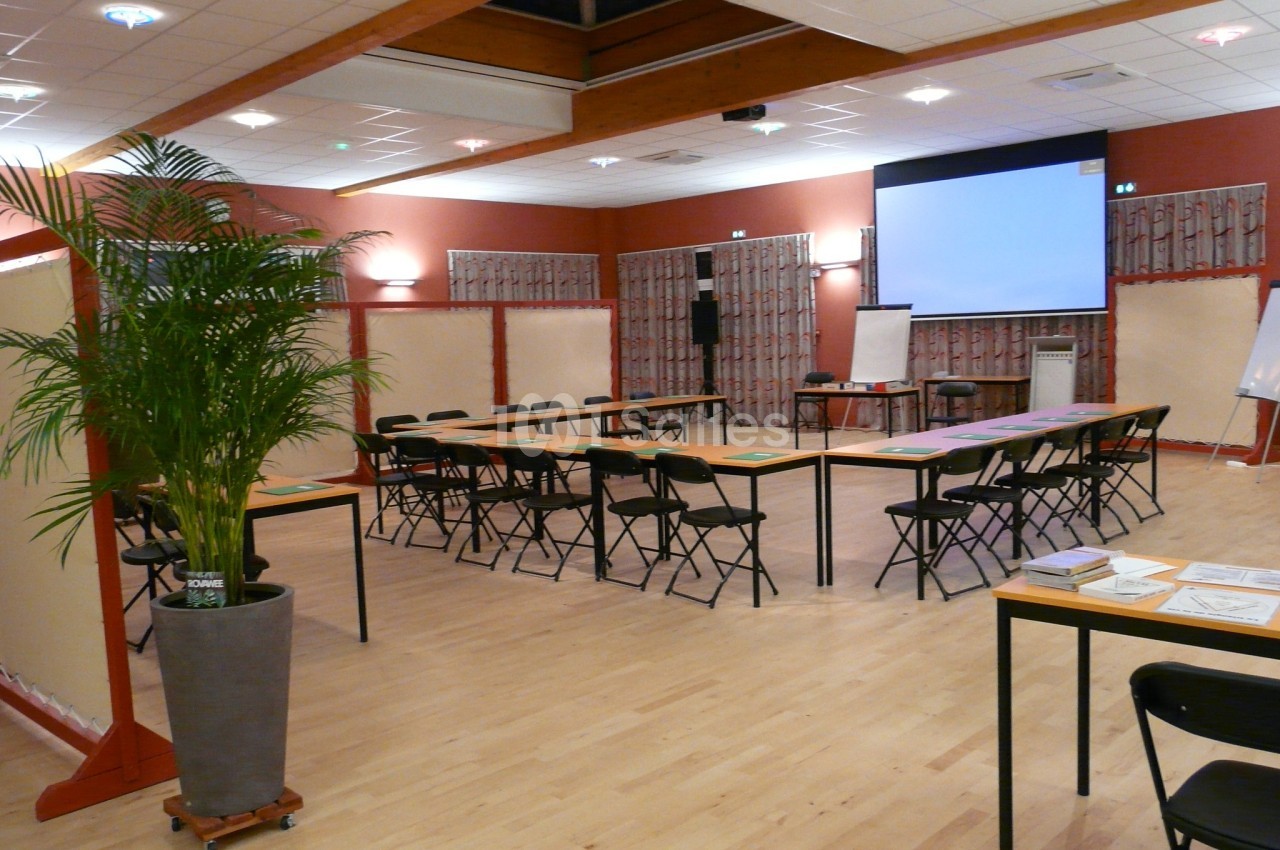 Salle de réunion avec tables disposées en U, chaises pliantes, écran de projection et plantes décoratives.