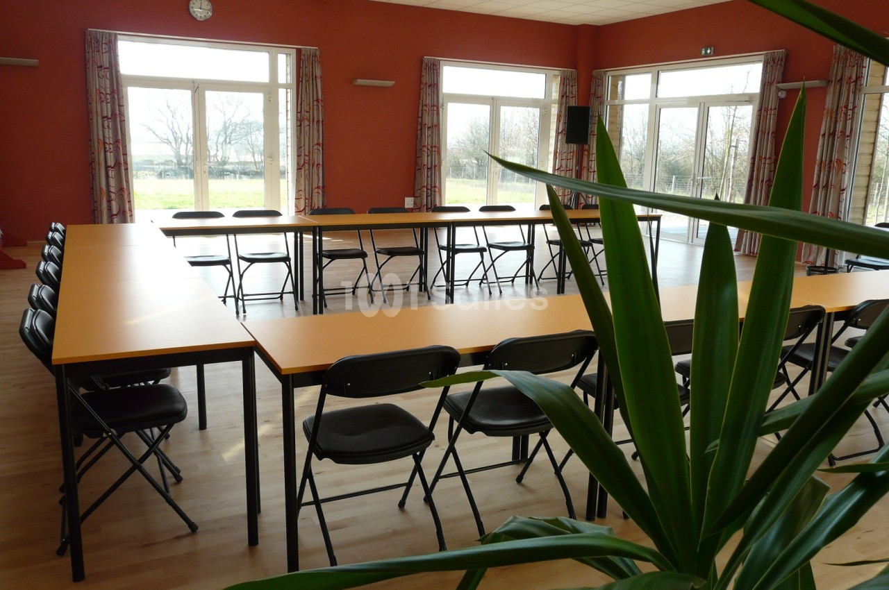 Salle lumineuse avec des tables disposées en U, des chaises pliantes noires et une plante au premier plan.