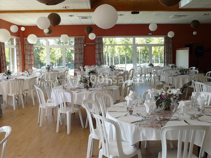 Salle décorée pour un événement, avec des tables rondes dressées, des chaises blanches et des lanternes suspendues.