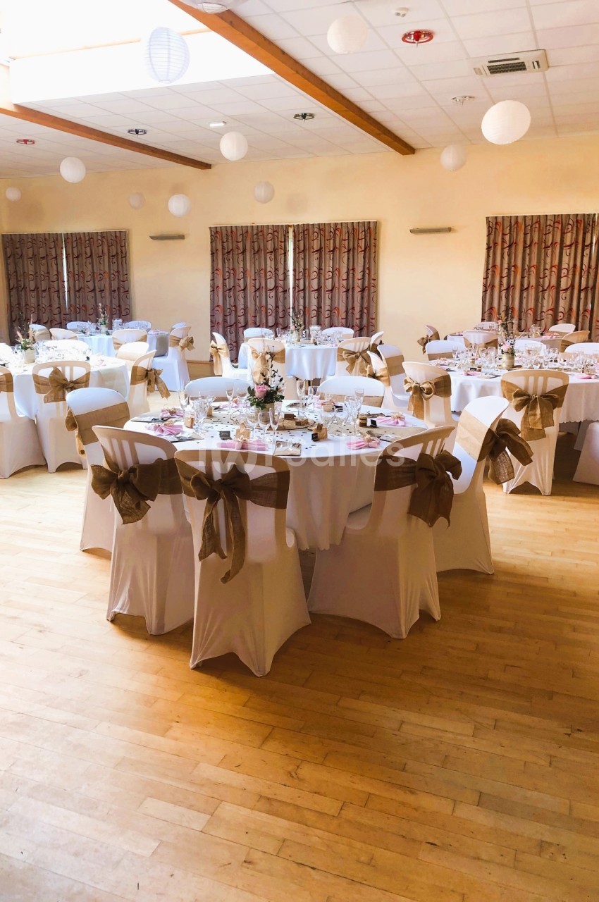 Salle décorée pour un événement avec des tables rondes dressées, nappes blanches et chaises ornées de nœuds beiges.