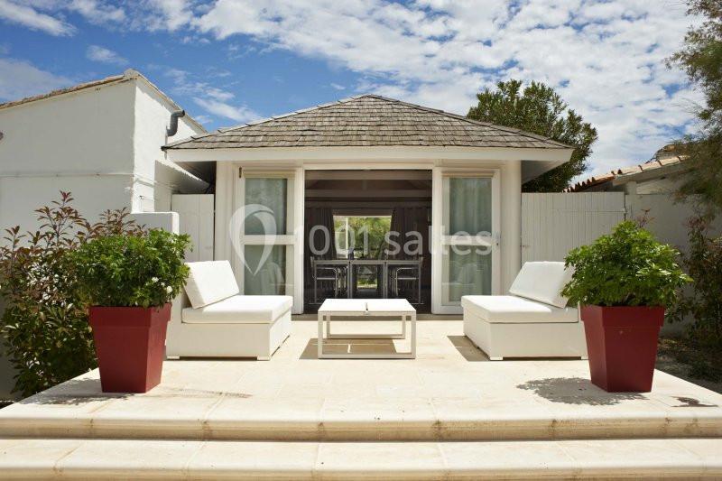 Terrasse avec deux fauteuils blancs, une table basse et des plantes en pots rouges devant une petite maison.
