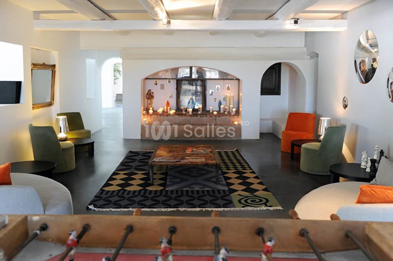 Salon lumineux avec canapés, fauteuils colorés, tapis à motifs, baby-foot au premier plan et bougies décoratives sur une…