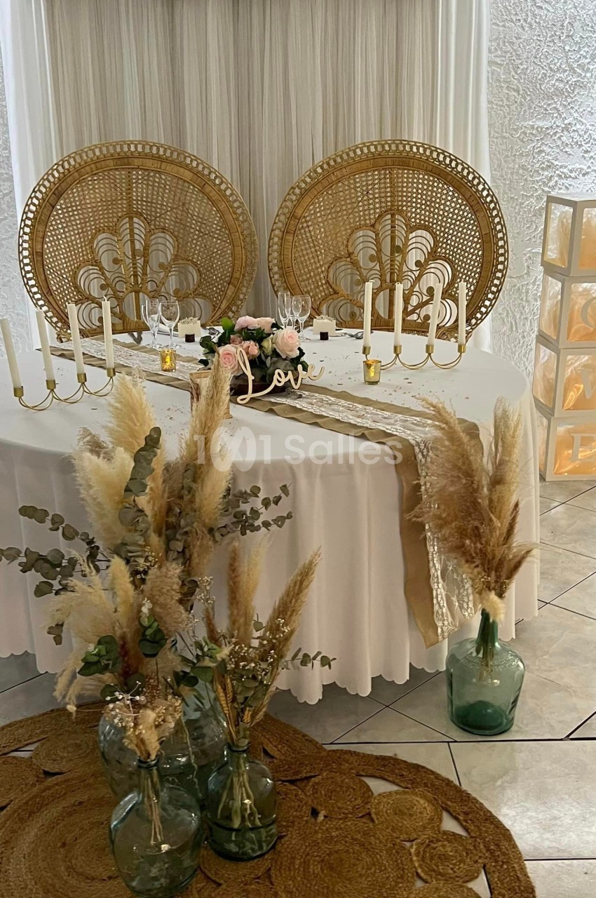 Table décorée pour un événement avec chandeliers, fleurs séchées, nappes blanches et chaises en rotin sculpté.